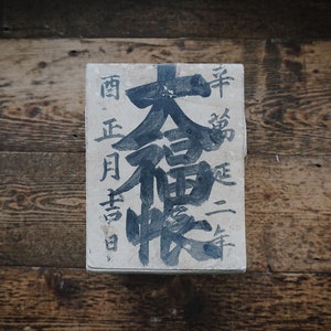May include: A white wooden sign with black Japanese calligraphy. The text reads "南 正月 吉日 大 福 壽", which translates to "South, New Year's Day, Auspicious Day, Great Fortune, Longevity".
