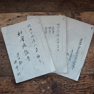 May include: Three pieces of old, white paper with black ink writing in Japanese. The paper is folded and stacked on a wooden surface.