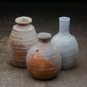 May include: Three ceramic vases of different shapes and sizes. The vase on the left is a light brown colour with a textured surface. The vase in the centre is a reddish-brown colour with a smooth surface. The vase on the right is a light grey colour with a textured surface.