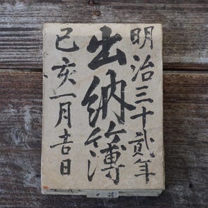 May include: A white paper sign with black Japanese characters. The text reads "明 治 三 十 年 亥 月 吉 日 生まれ", which translates to "Born on a good day of the tenth month of the year Meiji 30 (1897)".