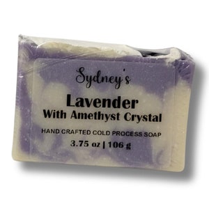 May include: A bar of handmade soap wrapped in clear plastic. The soap is lavender-colored with white swirls and includes the text "Sydney's Lavender With Amethyst Crystal." It weighs 3.75 oz (106 g).