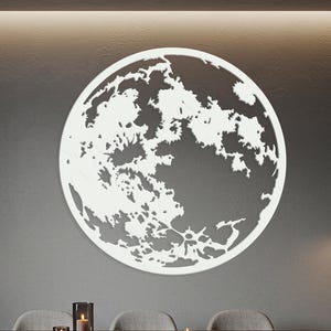 May include: A large, white, circular wall art piece depicting the moon's surface. The artwork is mounted on a gray wall above a dining table with chairs. The moon's craters and features are intricately cut out.
