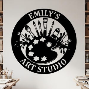 May include: Black and white wall art featuring a circular design with the text "EMILY'S ART STUDIO". The design includes paintbrushes, a paint palette, and a tube of paint, perfect for an art studio or creative space.