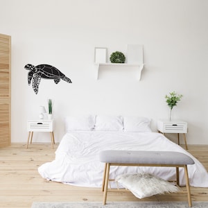 May include: A black sea turtle wall decal is mounted on a white wall above a bed. The bedroom features a white bedspread, two white nightstands, and a gray bench. The room has a minimalist, modern aesthetic with wooden floors.