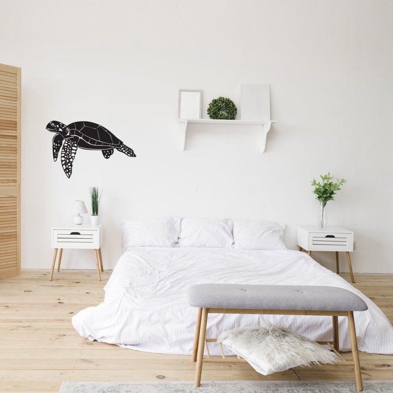 May include: A black sea turtle wall decal is mounted on a white wall above a bed. The bedroom features a white bedspread, two white nightstands, and a gray bench. The room has a minimalist, modern aesthetic with wooden floors.