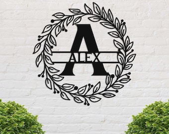 May include: Black metal wall art featuring the letter "A" with the name "ALEX" in the center. The letter is surrounded by a wreath of leaves and berries. The art is mounted on a white brick wall.