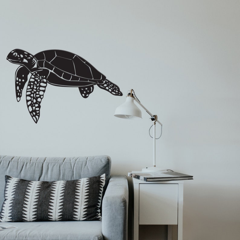 May include: Black sea turtle wall art. The wall art is a silhouette of a sea turtle with detailed flippers and shell. The turtle is depicted in a swimming pose. The art is mounted on a white wall above a gray sofa.