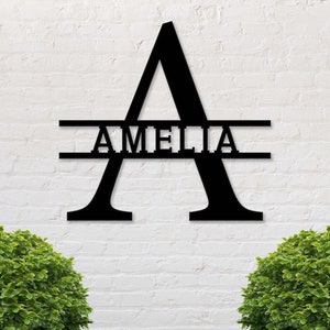 Puede incluir: Arte mural de metal negro con la letra "A" y el nombre "AMELIA" recortado en el centro. La obra de arte está montada en una pared de ladrillo blanco, con arbustos verdes en la parte inferior.