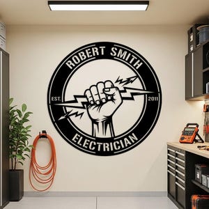 Puede incluir: Letrero de metal negro con un diseño circular que presenta un puño cerrado sosteniendo un rayo. El letrero incluye el nombre "ROBERT SMITH", la palabra "ELECTRICIAN" y los años "EST. 2011". El letrero está montado en una pared de color claro.