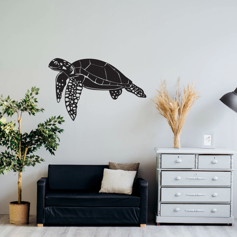 May include: A black sea turtle wall art decal. The turtle is depicted in a swimming pose, with detailed patterns on its flippers and shell. The decal is mounted on a light gray wall, creating a striking contrast. The room also features a black sofa, a dresser, and decorative plants.