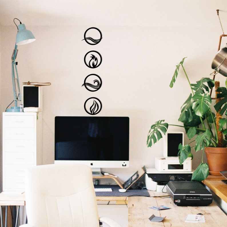 May include: Four black circular wall art pieces depicting the elements: water, fire, air, and earth. The minimalist designs are mounted on a white wall above a computer monitor. The room has a desk, chair, and a large potted plant.