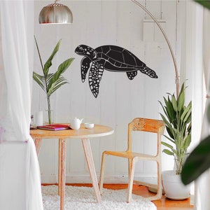 May include: A black metal sea turtle wall art decor. The turtle is depicted in a side profile, with detailed flippers and shell. The art is mounted on a white wall, creating a striking contrast. The room also features a wooden table and chair.