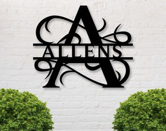 Peut inclure: Décoration murale en métal noir représentant la lettre "A" avec le nom "ALLENS" découpé. Le design comprend des volutes décoratives. L'œuvre est fixée sur un mur de briques blanches, avec des buissons verts en bas.