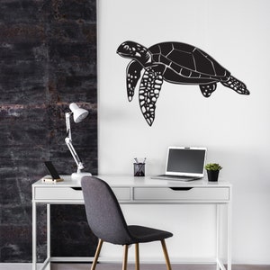 May include: A black metal sea turtle wall art decor. The turtle is depicted in a side profile, with detailed flippers and shell. The art is mounted on a white wall above a white desk with a laptop, lamp, and office supplies.