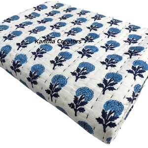 May include: A folded white quilt featuring a repeating pattern of blue floral motifs and dark blue stems. The fabric displays visible running stitch lines across the surface, and the text Kantha Creators is printed in black on the top section.