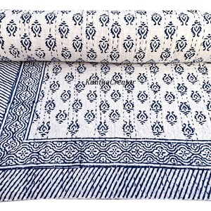 May include: A rolled-up white and navy blue quilt with a repeating floral pattern. The quilt is unfolded, revealing a larger section with the same pattern and a striped border. The quilt is made of soft material.
