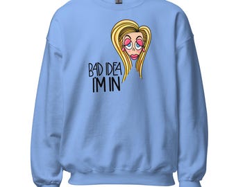 Può includere: Una felpa girocollo azzurra con un'illustrazione a cartone animato di una persona bionda con ombretto rosa e labbra grandi, posizionata sopra il testo nero "BAD IDEA I'M IN" scritto con un carattere in stile disegnato a mano sul davanti.