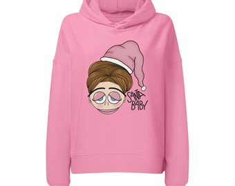 Può includere: Una felpa con cappuccio rosa acceso caratterizzata da un'illustrazione grafica di un volto con capelli castani che indossa un cappello da Babbo Natale rosa. L'illustrazione include il testo SANTA BABY in nero accanto al volto dagli occhi grandi.