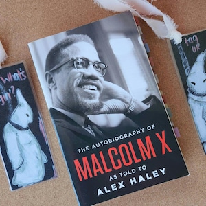 May include: A black and white photograph of Malcolm X on the cover of a book titled "The Autobiography of Malcolm X as told to Alex Haley". Three bookmarks are visible, one with a drawing of a ghost and the text "tag ur it", another with a drawing of a ghost and the text "What's Hangin?", and the third is a plain black bookmark.