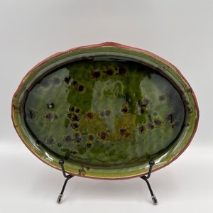 May include: Oval ceramic serving platter with a glossy green interior and a reddish-brown rim. The platter has a textured surface with dark brown spots and is displayed on a black metal stand. A decorative piece for serving food.