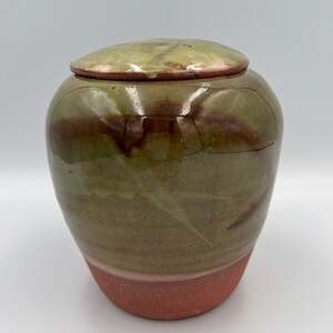 May include: A handmade ceramic jar with a lid. The jar has a rounded form, with a reddish-brown base, blending into green and brown hues. The lid is a matching green with brown detailing.