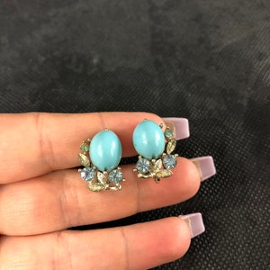 May include: A pair of clip-on earrings with a light blue oval-shaped stone surrounded by smaller clear and green stones. The earrings are made of gold-toned metal.