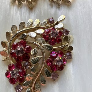 May include: A gold-tone metal brooch and matching pair of earrings featuring textured leaf motifs and clusters of faceted red and pink crystals. The floral-inspired jewelry set displays a polished finish with intricate detailing on the metal leaves.