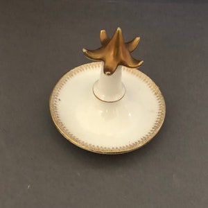 Puede incluir: Un porta anillos de porcelana blanca con un dise&ntilde;o pintado en oro. El porta anillos tiene una parte superior en forma de estrella elevada.