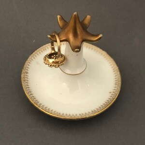 Puede incluir: Un porta anillos de porcelana blanca con un borde dorado y un soporte de anillo en forma de p&aacute;jaro dorado. Un anillo dorado est&aacute; en el soporte.