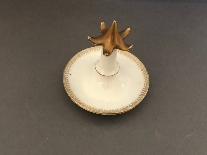 Puede incluir: Un porta anillos de porcelana blanca con un dise&ntilde;o pintado en oro. El porta anillos tiene una parte superior en forma de estrella elevada.