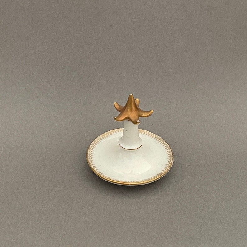 Puede incluir: Un porta anillos de porcelana blanca con la parte superior pintada en oro y un borde dorado. El porta anillos est&aacute; dise&ntilde;ado para sostener un anillo en la parte superior.