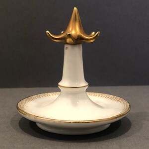 Puede incluir: Porta anillos de porcelana blanca con parte superior y base doradas. La parte superior tiene forma de flor con cinco p&eacute;talos.