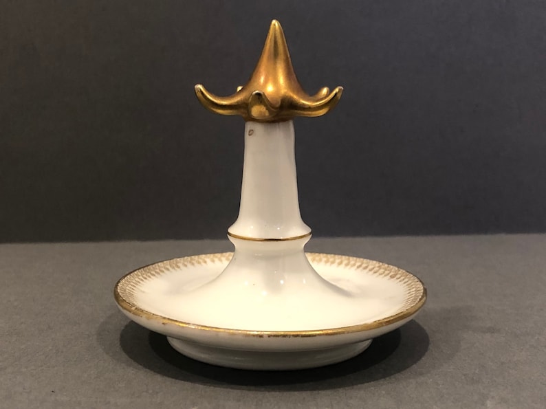 Puede incluir: Porta anillos de porcelana blanca con parte superior y base doradas. La parte superior tiene forma de flor con cinco p&eacute;talos.