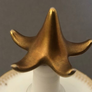 Puede incluir: Un remate de cer&aacute;mica en forma de estrella de cinco puntas de color dorado. El remate est&aacute; fijado a una base de cer&aacute;mica blanca.