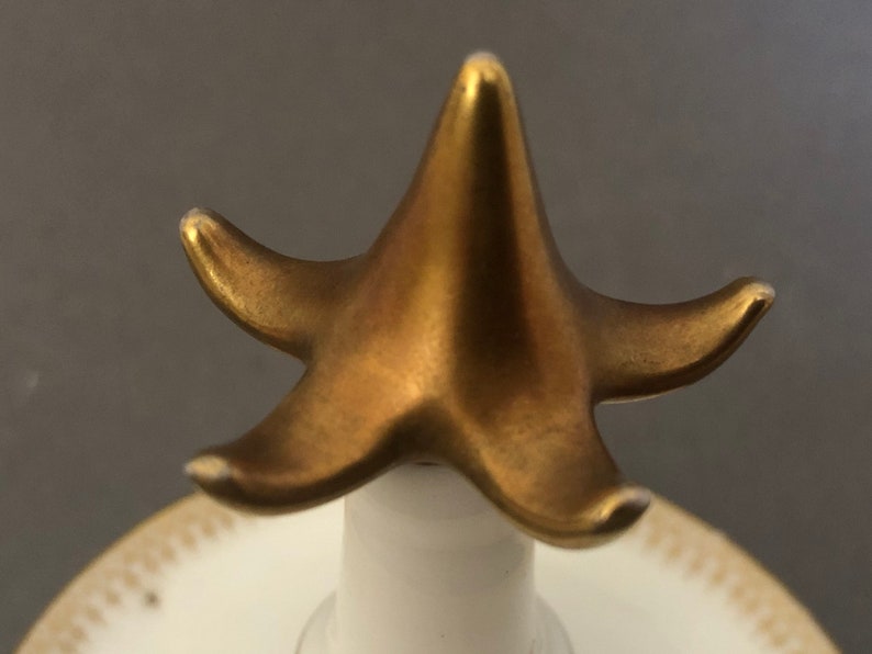 Puede incluir: Un remate de cer&aacute;mica en forma de estrella de cinco puntas de color dorado. El remate est&aacute; fijado a una base de cer&aacute;mica blanca.
