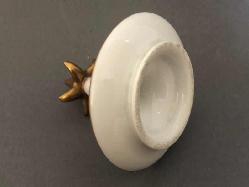 Puede incluir: Tapa de porcelana blanca con un pomo dorado en forma de estrella. La tapa es redonda y tiene una superficie lisa.