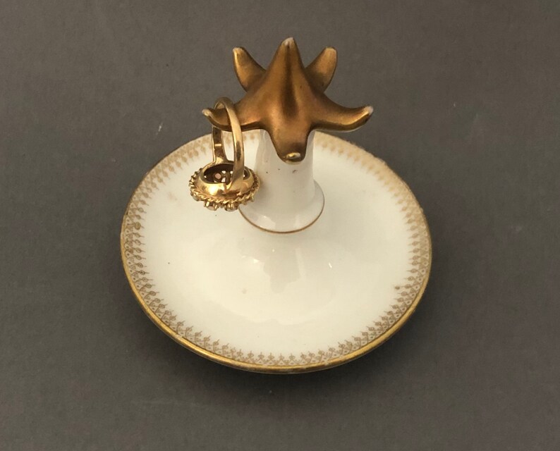 Puede incluir: Un porta anillos de porcelana blanca con un borde dorado y una parte superior en forma de p&aacute;jaro dorado. El porta anillos est&aacute; sobre un fondo gris.