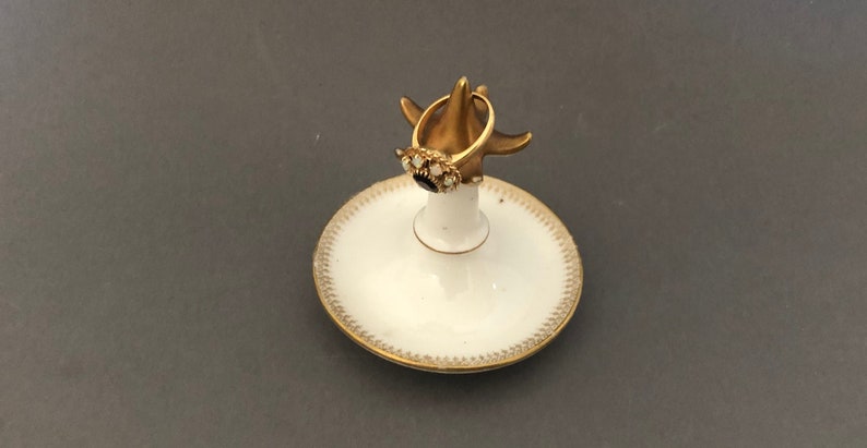 Puede incluir: Un porta anillos de porcelana blanca con un borde dorado y un soporte de anillo de oro. El soporte de anillo tiene un anillo con una piedra negra y piedras blancas.