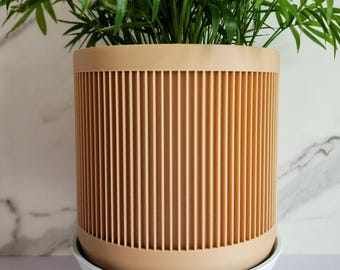 May include: A cylindrical light tan planter with vertical ribbed detailing, resting on a white circular base supported by three short, tapered legs. A lush green palm plant with thin, feathery fronds grows out of the top of the planter.