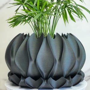 May include: A dark gray, decorative plant pot with a unique, petal-like design. The pot holds a vibrant green plant with long, slender leaves. The pot sits on a small, round, white table with wooden legs, against a marble background.