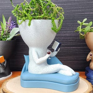 May include: Three decorative planters shaped like figures reading books and holding wine glasses. The planters are filled with plants and flowers. One figure is reclining on a blue chaise lounge, another is sitting on a blue chair, and the third is lying down.