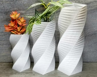 Może przedstawiać: Trzy białe ceramiczne wazony o spiralnym, fakturowanym wzorze. Wazony są ustawione w rzędzie od najmniejszego do największego. Najmniejszy wazon zawiera małą pomarańczową sukulentną roślinę, wazon środkowy zawiera zieloną roślinę z białymi paskami, a największy wazon jest pusty.
