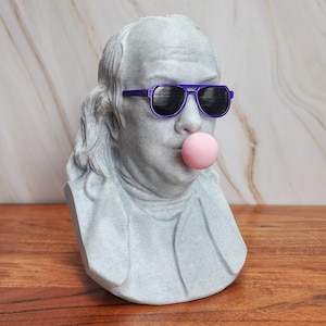 Puede incluir: Busto gris de Benjamin Franklin con gafas de sol moradas y soplando una burbuja rosa. La escultura está sobre una superficie de madera con un fondo de mármol. Un artículo decorativo único.