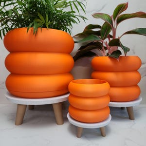 Pode incluir: Três vasos de anel empilhados laranja de vários tamanhos, cada um com uma planta diferente. Os vasos estão em suportes redondos brancos com pernas de madeira. As plantas têm folhas verdes e vermelhas.