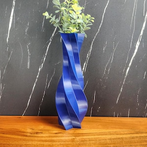 Puede incluir: Un jarrón de cerámica azul con un diseño retorcido. El jarrón está lleno de plantas verdes.