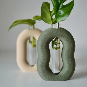 Może przedstawiać: Dwa dekoracyjne wazony o unikalnym, otwartym okrągłym kształcie. Jeden wazon jest w kolorze jasnobeżowym, a drugi w stonowanej zieleni. Każdy wazon zawiera przezroczystą szklaną tubę z zieloną łodygą i liściem rośliny. Wazony to nowoczesny element wystroju wnętrz.
