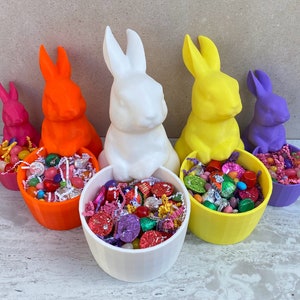 May include: Five colorful bunny figurines with candy-filled bowls. The bunnies are white, orange, yellow, purple, and light pink. The bowls are filled with colorful candies.