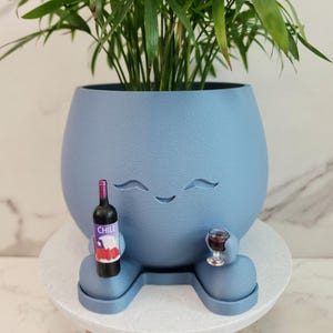 May include: A matte blue rounded planter with a smiling face, featuring two small arms and legs. The planter holds a green plant and is holding a miniature wine bottle with a label reading CHILE and a small glass of red wine in its hands.