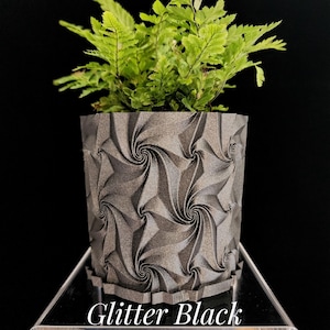 May include: A cylindrical planter with a dark gray glitter finish and a repeating geometric spiral pattern. A vibrant green fern plant is potted inside the container. The text "Glitter Black" is printed in white at the bottom of the image.