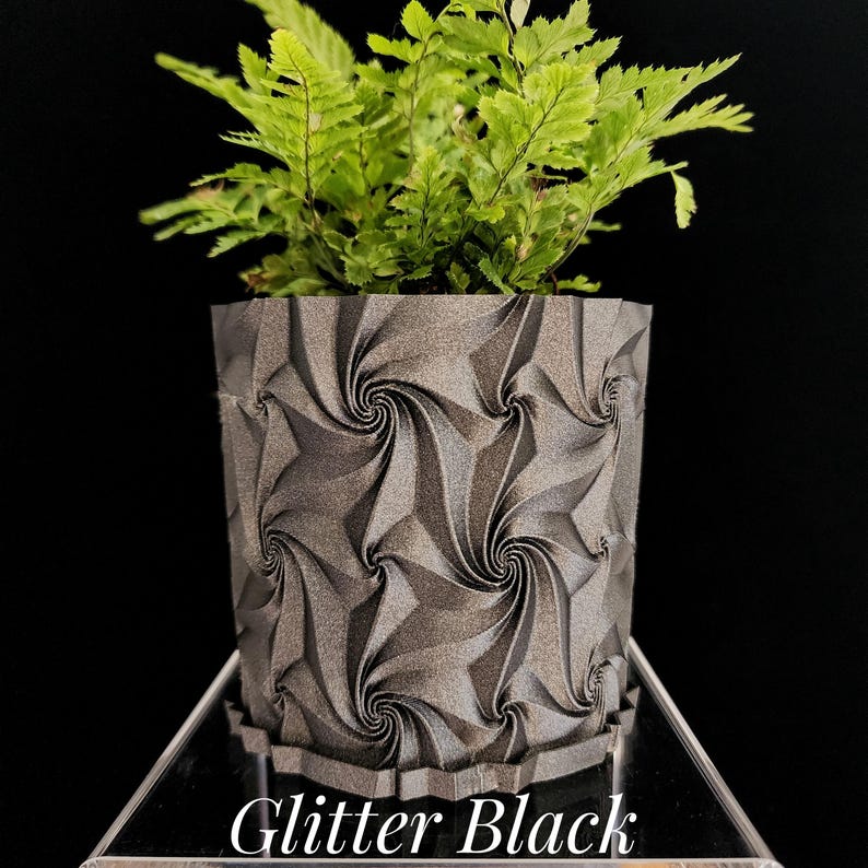 May include: A cylindrical planter with a dark gray glitter finish and a repeating geometric spiral pattern. A vibrant green fern plant is potted inside the container. The text "Glitter Black" is printed in white at the bottom of the image.